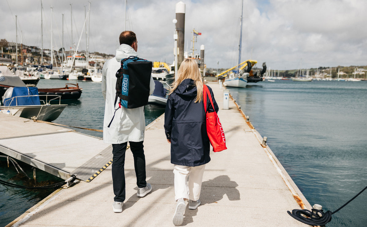 Two people walking away on a concrete dock at a marina, one wearing a white hooded jacket with a black backpack and the other wearing a navy hooded jacket and carrying a red bag.