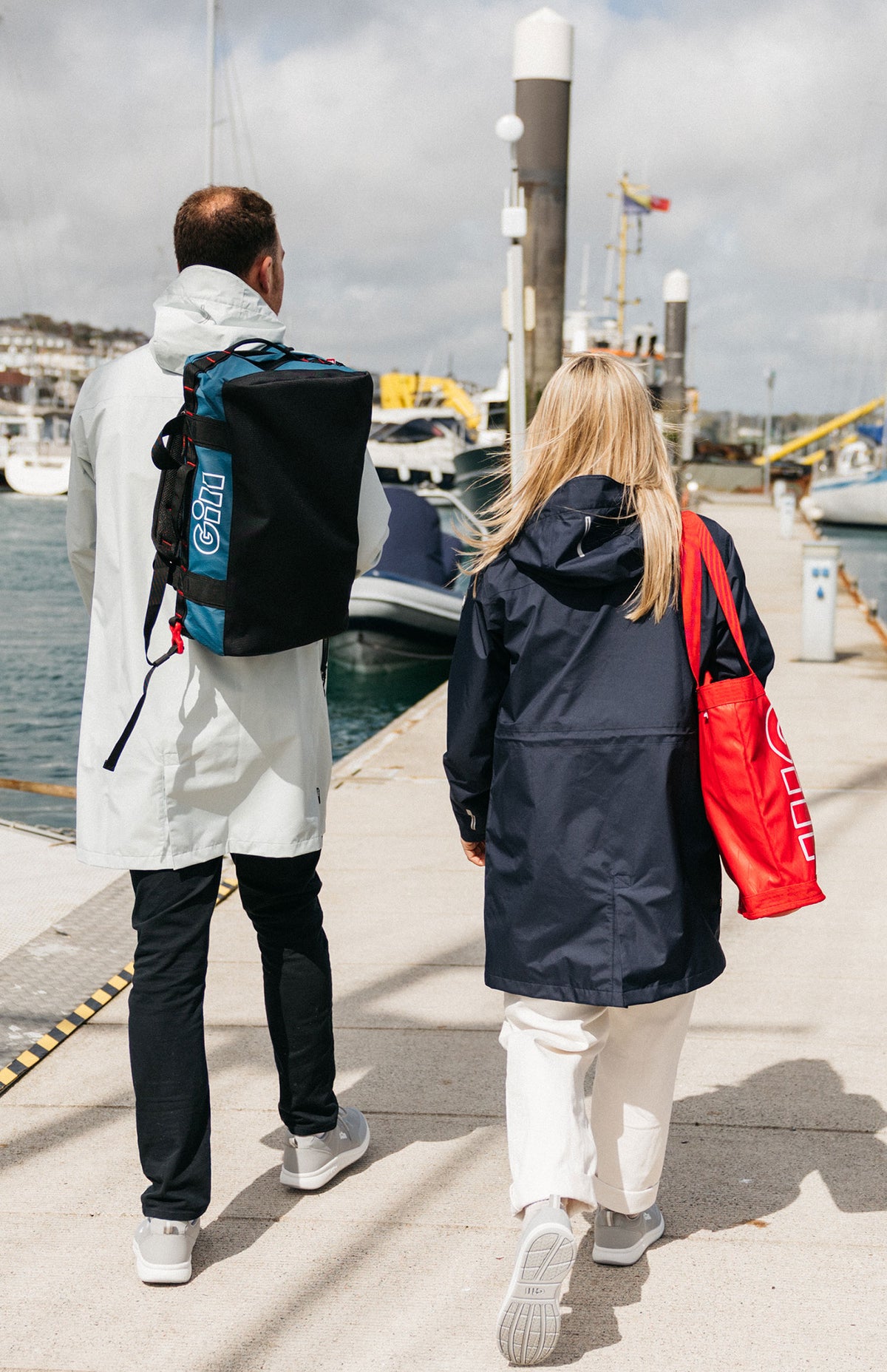 Two people walking away from the camera on a sunny day at a harbor, showcasing a black and blue GILL backpack worn by the man in a white jacket, and a woman in a navy jacket carrying a red tote bag.