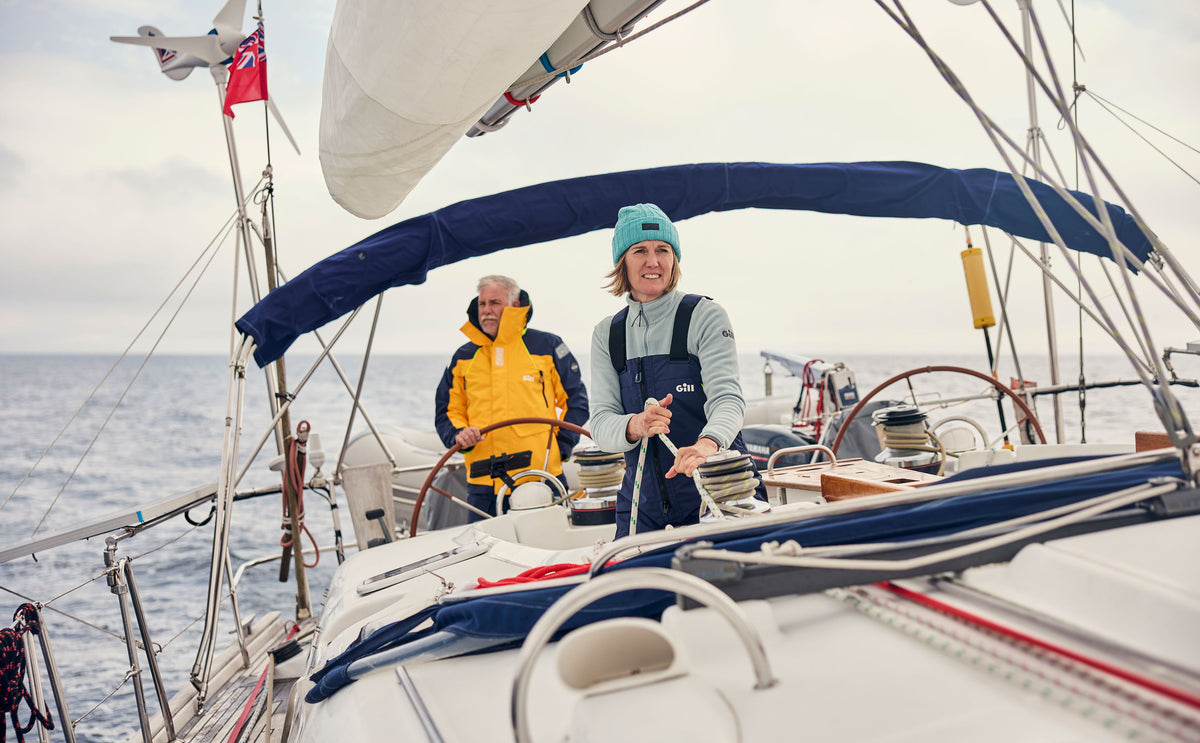 Two people sailing on a yacht in the open ocean under an overcast sky. The person in the foreground wears a light blue beanie and dark blue overalls, steering the boat, while another person in a yellow jacket sits behind them.