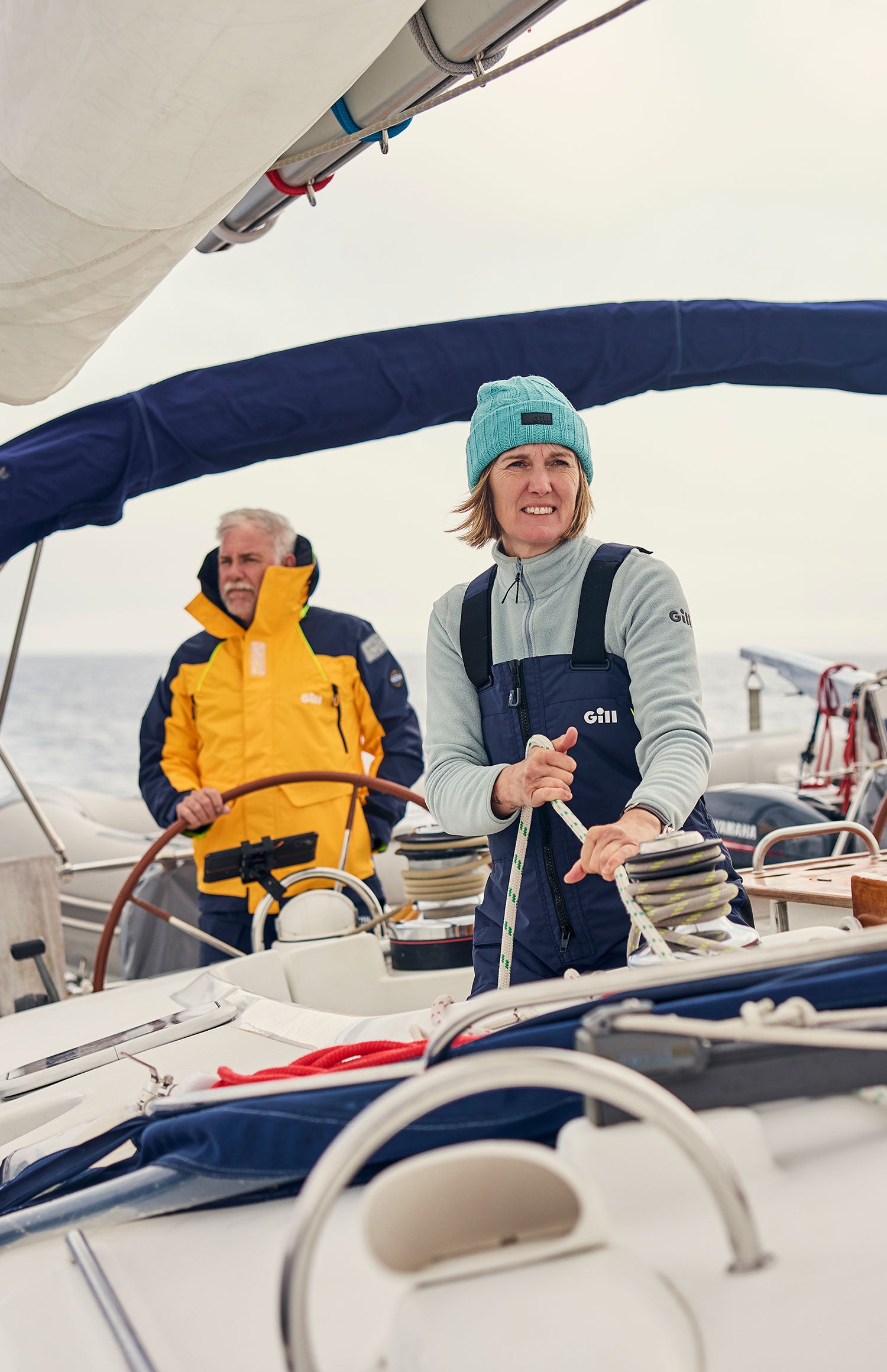 A woman in a teal beanie and navy overalls steering a sailboat, with an older man in a yellow and navy jacket standing behind her. The boat deck and rigging are visible, with an overcast sky in the background.