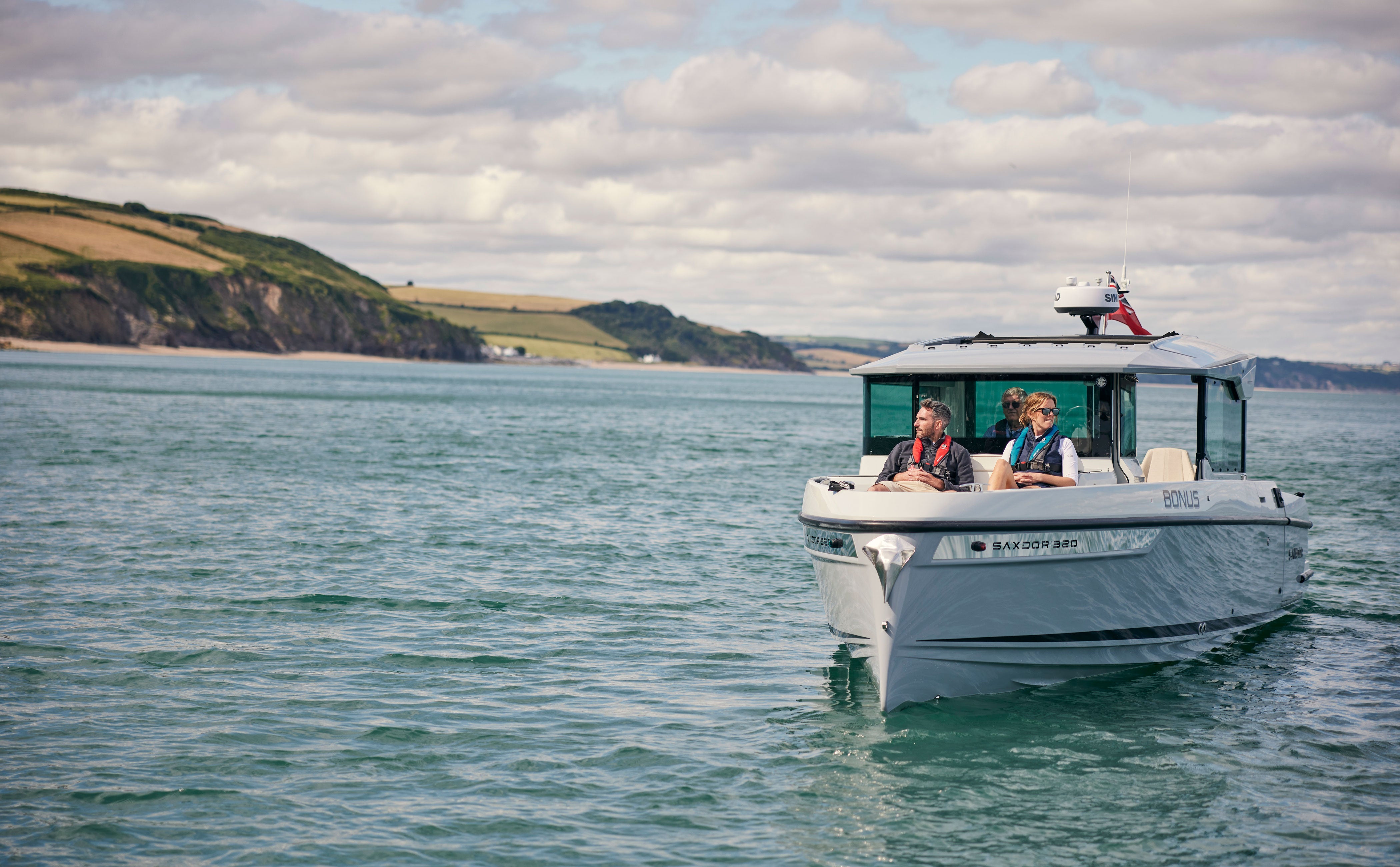 A white motorboat with two people on board sailing on blue-green water under a cloudy sky, with a coastal headland visible in the background.