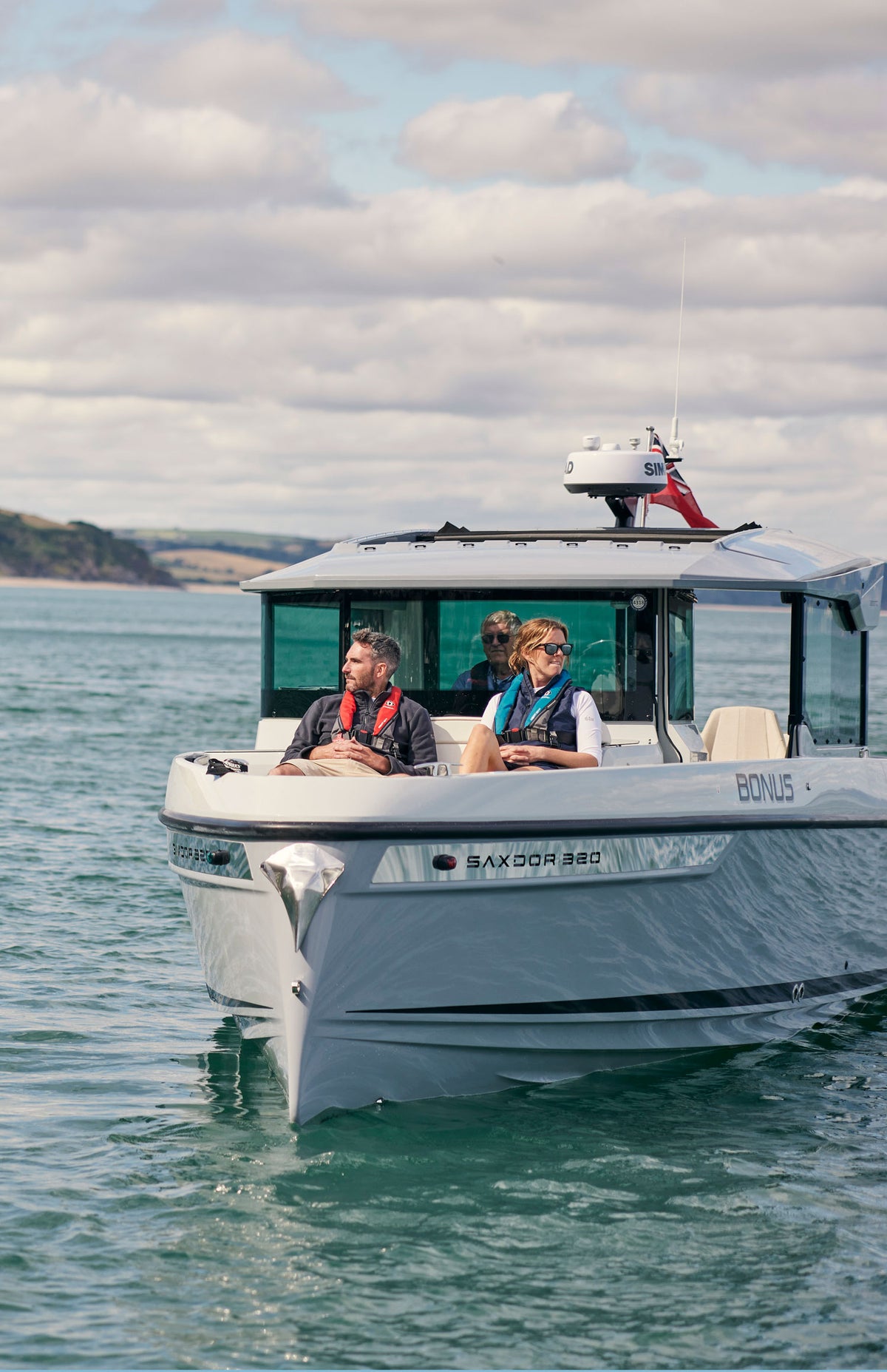 A white motorboat with two passengers cruising on blue-green water under a cloudy sky. The boat features a hardtop cabin, tinted windows, and a radar antenna on top, with the name 'BONUS' visible on the side.