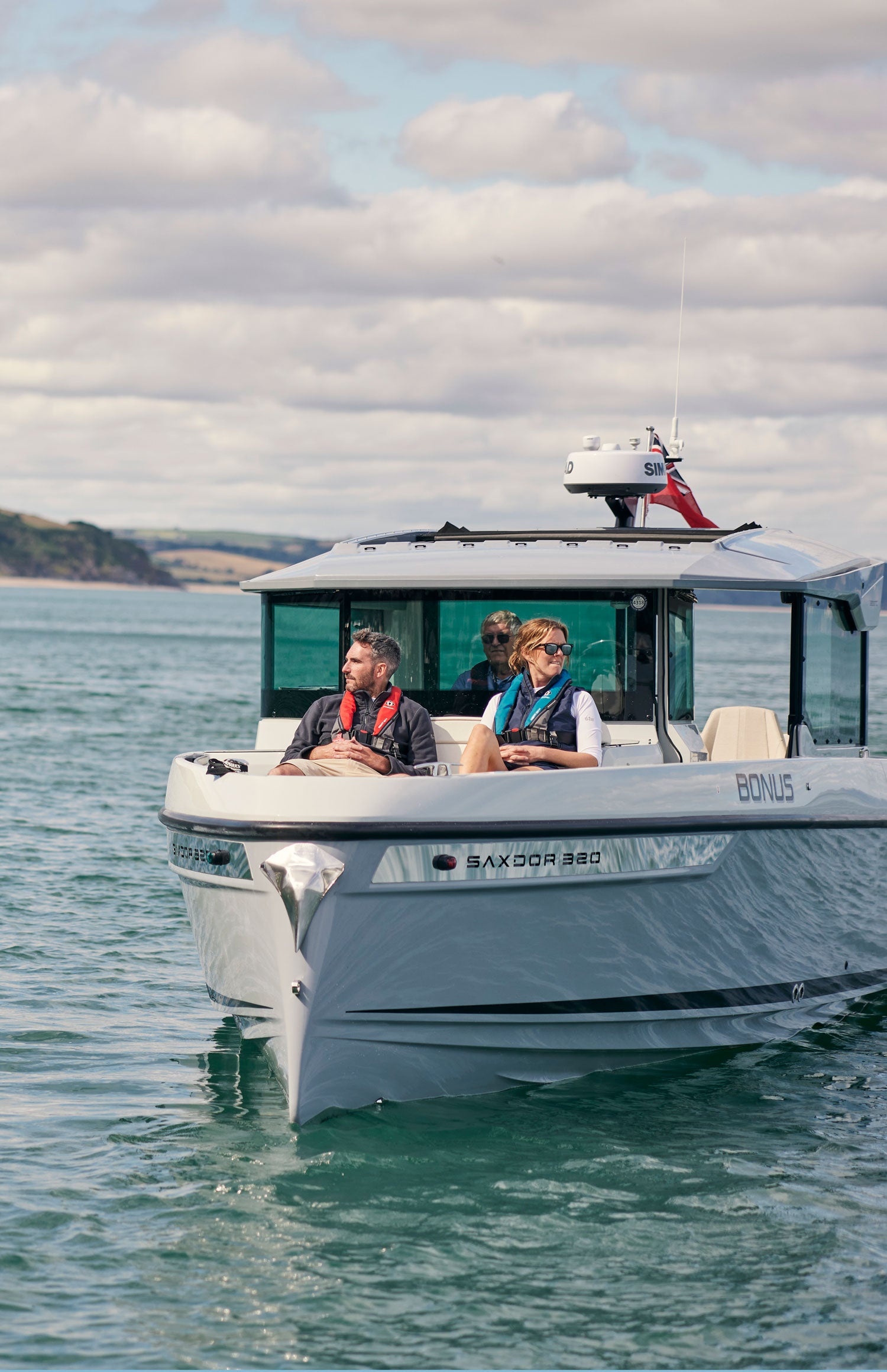 A white motorboat with two passengers cruising on blue-green water under a cloudy sky. The boat features a hardtop cabin, tinted windows, and a radar antenna on top, with the name 'BONUS' visible on the side.