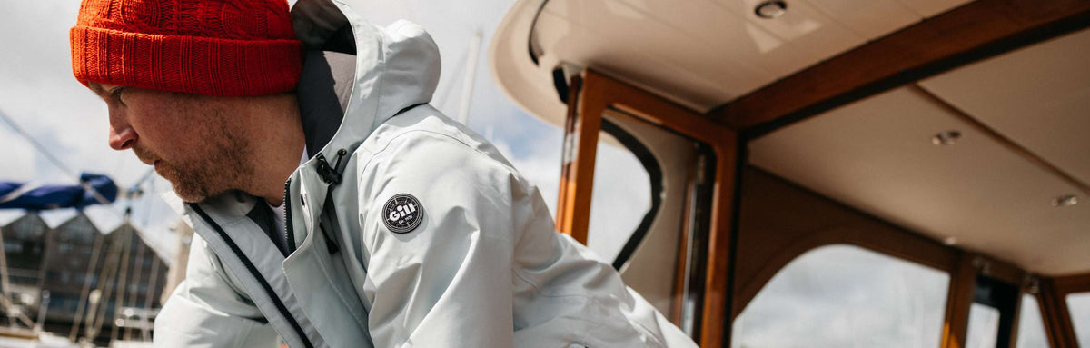 A man wearing a light gray hooded jacket and a bright red knit beanie sits inside a boat cabin. A circular Gill logo patch is visible on the sleeve of the jacket.