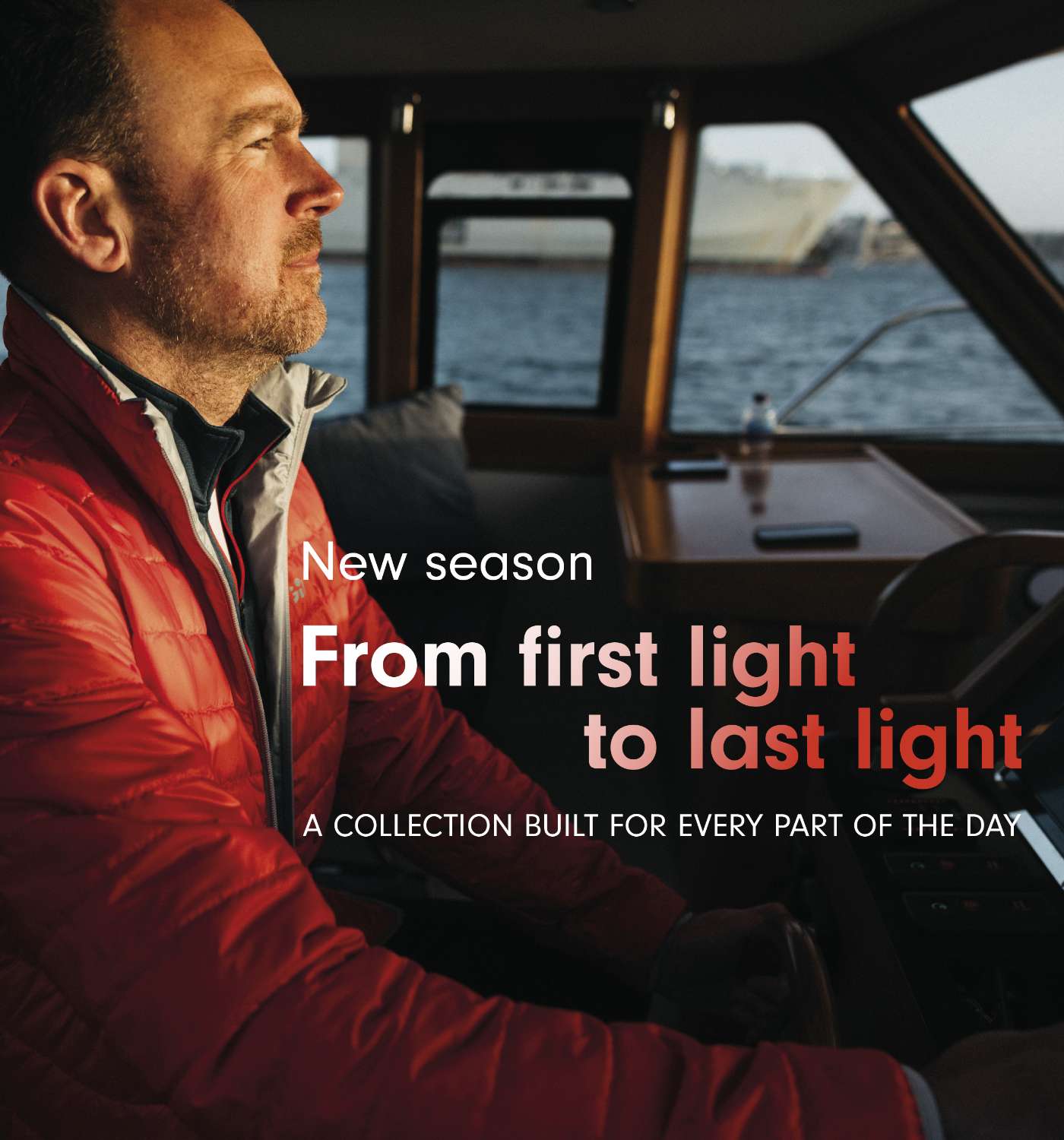 A man in a red puffer jacket sits inside a boat cabin, looking out toward the water. White and coral text overlays read, 'New season From first light to last light A COLLECTION BUILT FOR EVERY PART OF THE DAY.