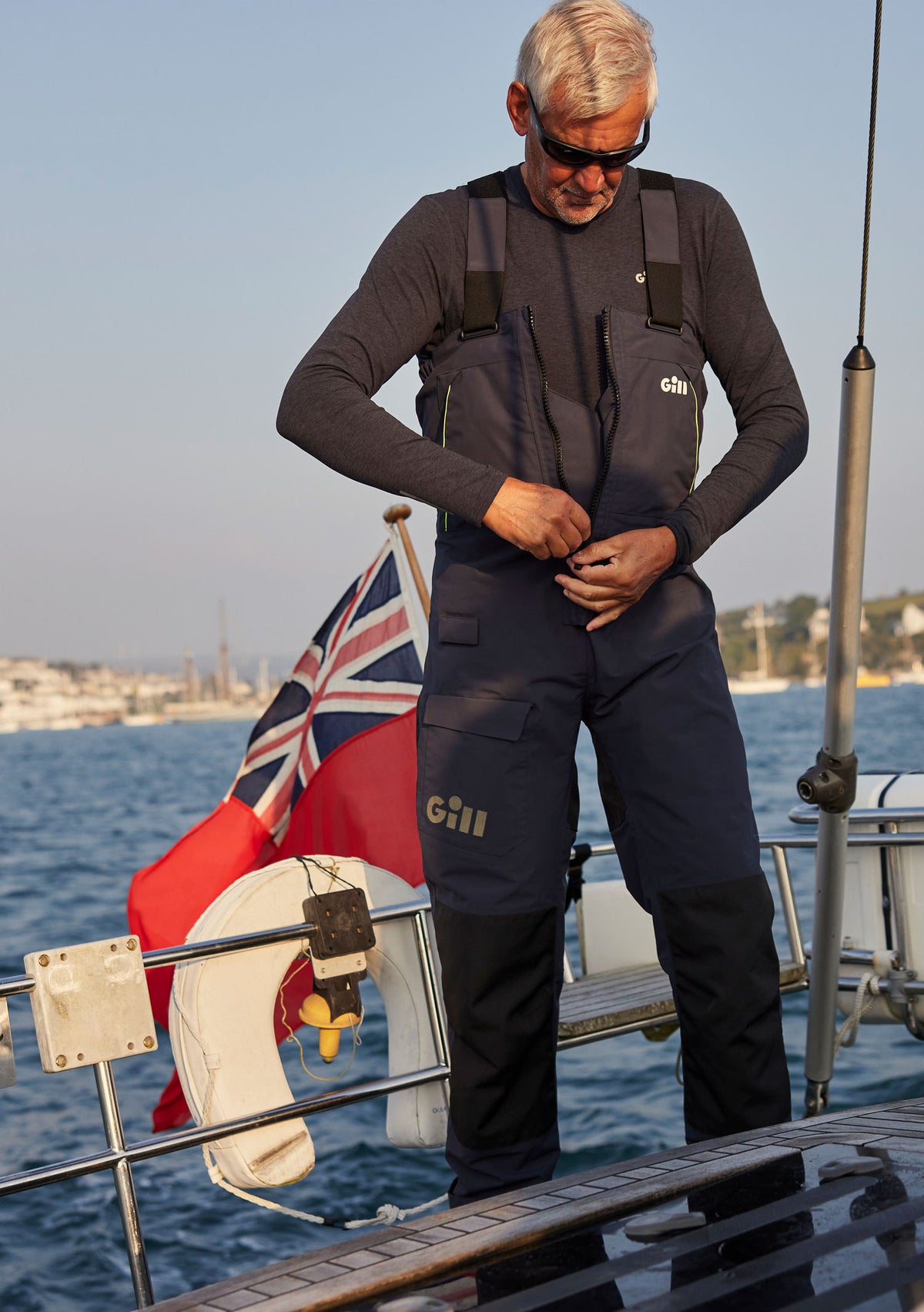 A man wearing a dark gray long-sleeve top and navy blue Gill brand waterproof overalls stands on a boat deck by the water, adjusting the front of the overalls. A Union Jack flag and white lifebuoy are visible in the background.