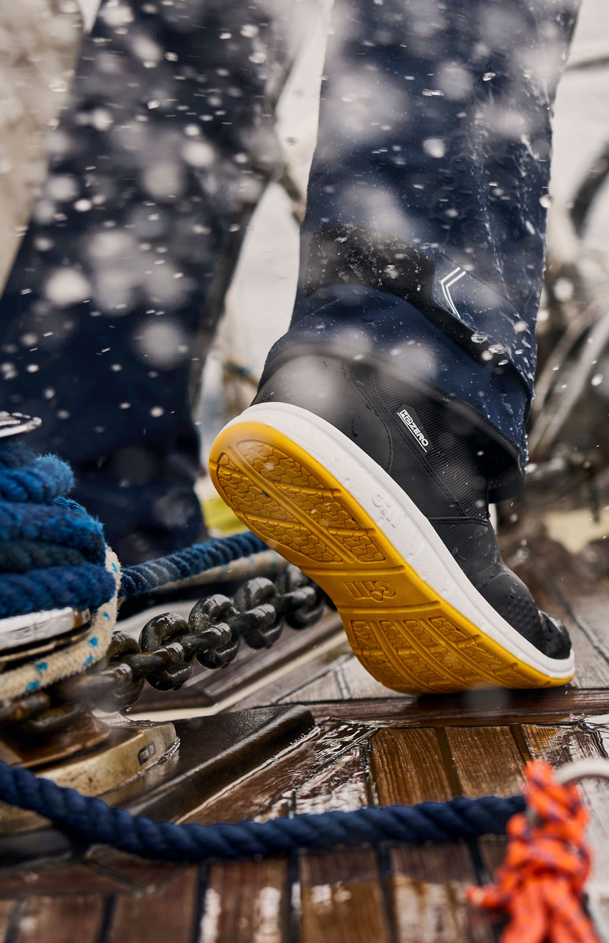 A close-up of a person's foot wearing a black waterproof shoe with a yellow sole, standing on a wet wooden deck with ropes and splashing water in the background.