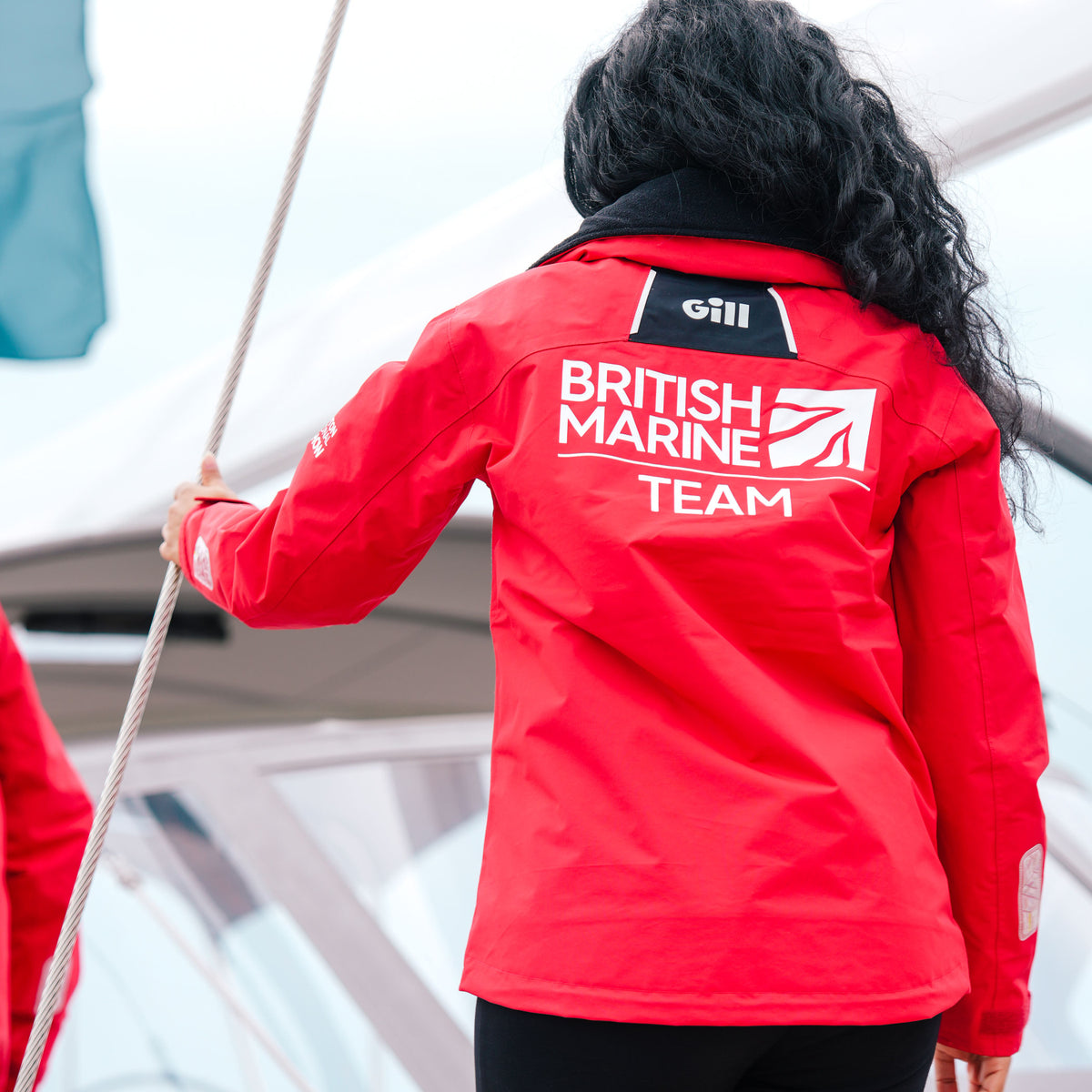 A person viewed from behind wearing a bright red sailing jacket with 'BRITISH MARINE TEAM' printed in white on the back, standing on a boat deck.