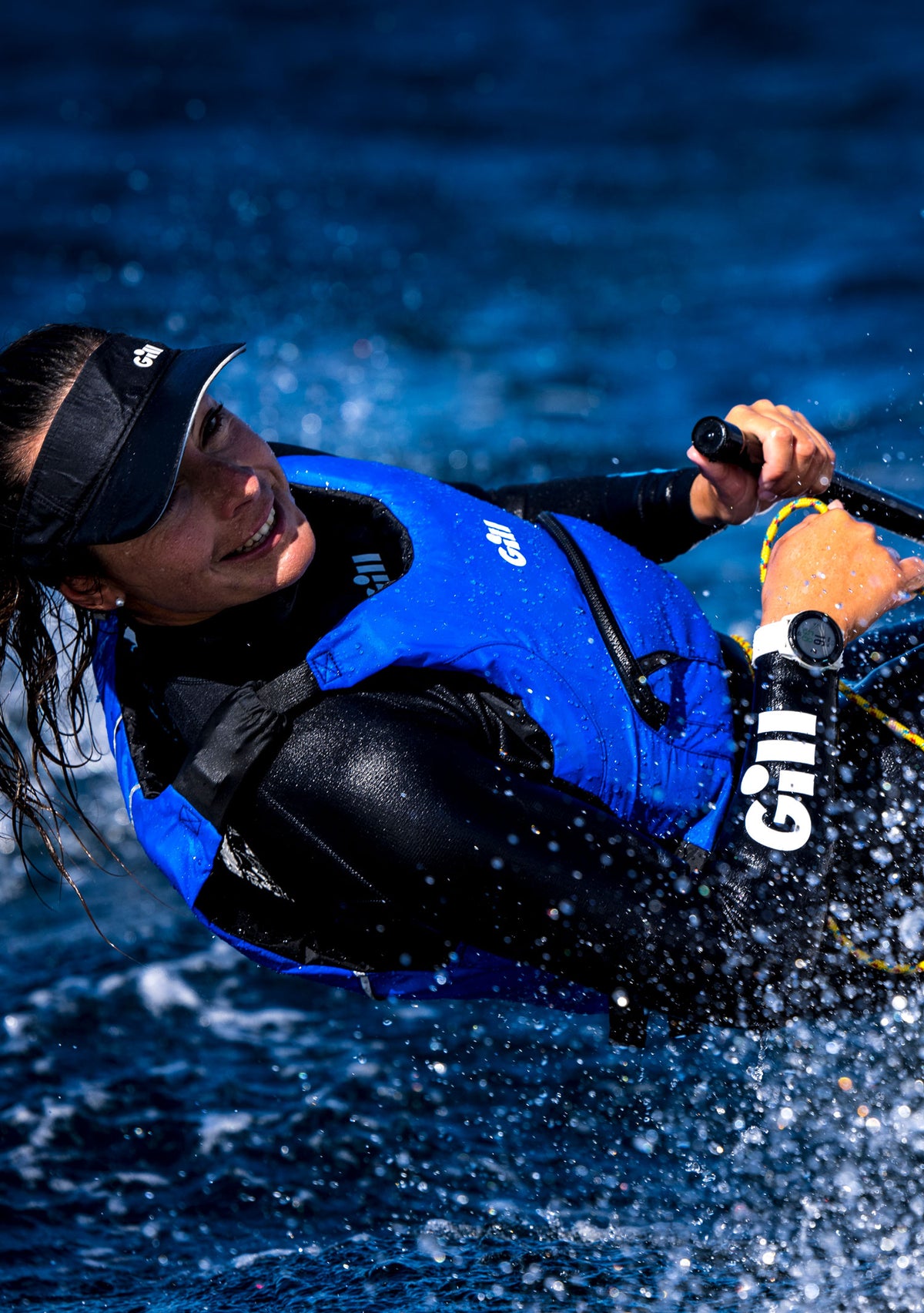 A woman in a black wetsuit and blue life vest riding on a paddleboard in deep blue water, with water splashing around her. She is wearing a black visor and a watch.